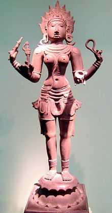 A reddish bronze statue of a four-armed topless woman wearing a short pant sari, standing on a pedestal. She wears a flaming crown and various ornaments. She carries in her hands a goad, a noose, a bowl and a small broken trident.