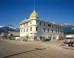 alt=Photograph of the Golden North Hotel in the Skagway Historic District, and other historic buildings, across a broad, unbusy street with dramatic mountains behind.