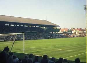 A colour photograph, taken from behind a goal net, of a roofed English-style grandstand, filled with people