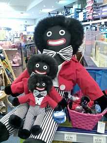 A rag doll, his face a caricature of a black person, in a toy store, holding copies of himself in his lap and more copies in a basket.