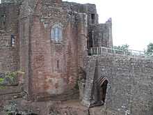 A castle, with a flat fronted tower facing the viewer with a stained glass window in the middle of it; a stone causeway is on the right of the picture, leading to a gateway to the right of the tower – a partially filled arch is supporting the causeway.