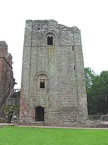 A square stone keep dominates the picture, sat behind a patch of green grass; the keep has a doorway at ground level, with two windows irregularly placed above it.