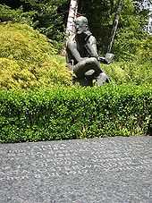 Horizontal gravestone saying "JAMES JOYCE", "NORA BARNACLE JOYCE", GEORGE JOYCE", and "...ASTA OSTERWALDER JO...", all with dates. Behind the stone is a green hedge and a seated statue of Joyce holding a book and pondering.