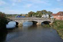 A stone three arch bridge over water. On the bridge is a small blue lorry. Either side of the river is vegetation and to the right of the bridge houses.