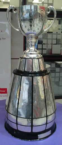 A trophy in the design of a silver cup affixed to a large, round wooden base.  The base has silver plates attached to it engraved with the names of previous winners.