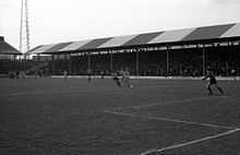 A black-and-white photograph of a football match in progress in front of a modest, single-tiered, British-style grandstand