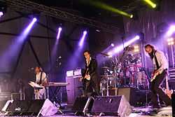 A rock band performing live on-stage. The stage is brightly lit On the left, a man plays bass behind a keyboard. In the centre, a man plays guitar and behind him a man plays drums. On the right, a man plays an electric mandolin.