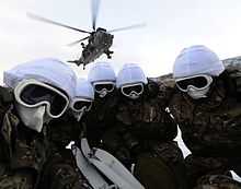 Colour photograph of five fully covered marines in a huddle underneath a helicopter.