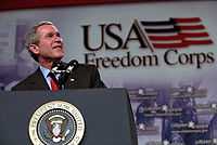 President George W. Bush stands at a podium with a large banner behind him that says “USA Freedom Corps”.