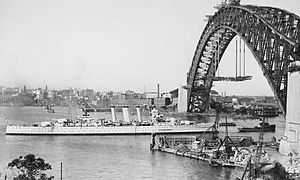 A black-and-white photograph of a ship sailing under a partially constructed bridge under a clear sky with the shore visible on the horizon