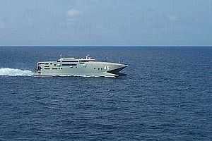 A warship-grey catamaran travelling at speed (from right to left) on the open sea