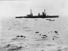 A large warship lowers boats to pick up sailors floating in the open water