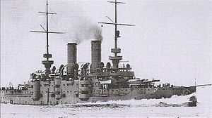 A small grey battleship traveling at full speed with dark smoke billowing out of its two funnels.