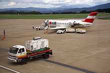 A Bombardier CRJ-200 aircraft parked on the tarmac with passengers approaching the aircraft for boarding. There is also a baggage trolley and two carts next to the aircraft. There is an auxiliary power unit in the foreground and a mountain view in the background