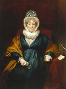 Oil painting of a woman sitting in a striped chair. She is wearing a dark-colored dress, with a shawl, contrasted with her tight, white cap and collar. Next to her is a table with writing instruments.