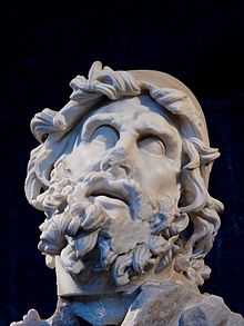  Stone image of the head of a man, showing signs of erosion due to age. The most striking feature is the heavily coiled beard and hair. The eyes are looking sightlessly upwards