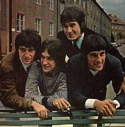 Four smiling young men leaning over the back of a green park bench, a row of three-story-tall residential buildings behind them. The man on the left wears a brown sports jacket and white turtleneck; the man to his left wears a black-and-white-striped pullover shirt; the man to his left (standing straighter, just behind the other three) wears a black suit and tie; the man on our far right wears a black sports jacket and white shirt.