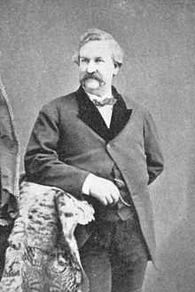 A monochrome photograph of a man from the thigh up, approximately 40 years old, with a large mustache and no sideburns, leaning with his right elbow on a fur-draped arm of furniture, looking slightly to the left