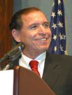 Head and shoulders of a man in his forties with close-cropped dark hair, dressed in a dark blue suit, a white shirt and a red striped tie. On his right lapel is the President’s “Call to Service” Award medal. Over his right shoulder the U.S. flag.
