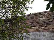 The bridge from the road, looking up through foliage at the east parapet wall with its unornamented date stone.