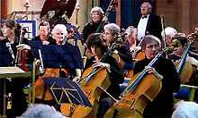 A selected view of an orchestra playing, cellos at the front, double basses behind.