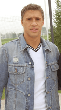 A man dressed in denim jacket and white t-shirt stands outside a football training pitch.