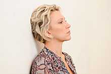 Hamann in profile with short, wavy blonde hair, chin raised and head against an pale ivory-colored wall