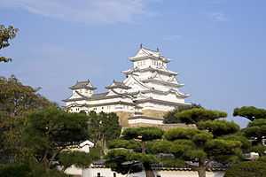 Photo of the central castle towers in the Himeji Castle complex, projected against the blue sky. Trees can be seen in the grounds below the castle towers.