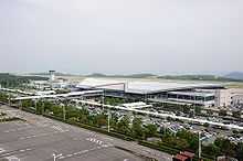 An airport terminal building with an air traffic control tower on the left and a carpark on the foreground of the building