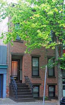 A narrow brick building with dark brown steps leading to the main entrance. Most of the upper two stories are obscured by a tree
