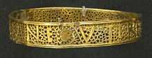 A gold bracelet with a pattern and writing. The upper and lower edges are solid, but between them is a lacey pattern made of leafy plant tendrils. Amidst this mostly perforated pattern, letters are formed from solid gold segments.