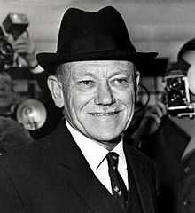 A middle-aged man wearing a dark suit and tie, and a hat. He has a toothbrush moustache.