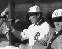 A man in a white baseball jersey and white baseball cap with a dark brim and an Old English "D" on the front points at something off-camera with his right hand.