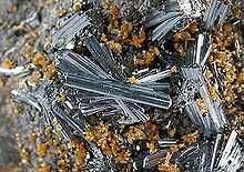 A close view of a rock crusted with groups of glassy, lustrous, silvery-blue hutchinsonite, in tight clusters of loosely aligned needle-like crystals, among smaller clusters of tiny orange-brown crystals