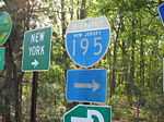 A grouping of road signs, with two fully visible. The left sign is a green square that reads New York right arrow and the right sign is an old shield that reads Interstate 195 right.