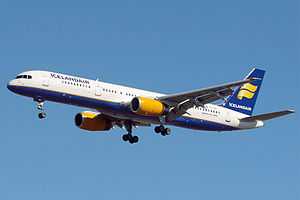 A mostly white Boeing 757 with blue and yellow trim preparing for landing against a blue sky. Landing gear and flaps are extended in final approach configuration.