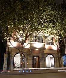 photo of a colonial style building lit up at night. The first two floors of the building are visible, with the top floors obscured by trees with dense foliage. The interior of the gallery can be viewed from outside through two ceiling to floor windows.