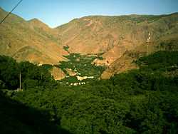 The village of Imlil in its wooded valley, surround by arid mountains. Taken early on a summer's morning, facing west. To the right a mobile phone mast sits incongruously on a nearby hill. Terracing of the valley is visible behind the village. Tall green trees are in the foreground.