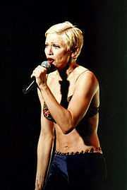 Image of a blond female facing her left. She has short blonde hair and is wearing a green bra and purple pants with beads on the waist. She is singing to a microphone, which she holds to her mouth with her left arm.