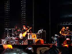 Two guitarists, a drummer and a keyboard player are performing on an orange lit stage in front of an audience. Various pieces of equipment surround the group. The stage features a black backdrop with two strips of vertical lights.