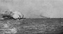 A ball of flame engulfs a large gray warship. Several smaller ships are seen in the distance.