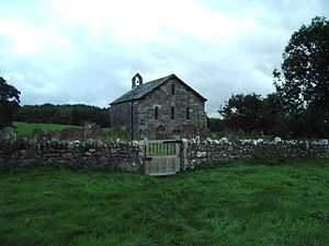 A small, simple, stone church, in front of which is a stone wall.  On the far gable is a bellcote with a single bell.