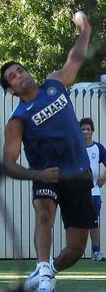 Young brown skinned clean shaven man wearing a blue sleeveless T shirt with the word "SAHARA" in black, wearing dark shorts is about to deliver a white cricket ball. His left bowling arm is above his head and he is wearing white sports shoes. His leading right leg is straight while his trailing left leg is bent. He is grimacing. In the background is a white picket fence and shady green vegetation.