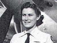 A woman in a neat, white uniform with a black tie with dark hair that is tied back stands at attention in front of an airplane and only the area from her shoulders up are visible in black and white. She is smiling.