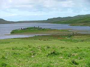 A small island in a lake lies offshore from green fields. A small wooden footbridge leads to the islet which contains various stone ruins including at least two gable ends.