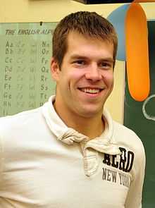 A man standing directly in front of the camera. He has short brown hair and is wearing a light colored polo shirt.