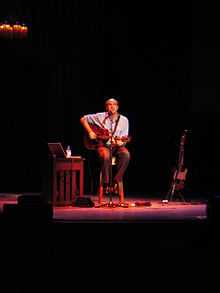 A man behind a microphone holding an acoustic guitar, sitting on a stool in the center of a stage, with a light shining down from above. To his sides are an electric guitar on a stand, a side table with a laptop, a bottle, and some additional equipment.
