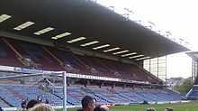 A two-tiered cantilever football stand. The lower tier has light blue seats with some claret seats which spell the word "Clarets". The upper has all claret seating with light blue seats spelling "Burnley".