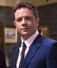 Head and shoulders photograph taken indoors of a man with short, spiky hair wearing a suit collar and tie, looking direct to camera. A microphone is attached to his tie.