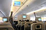 Airliner cabin. Rows of seats arranged between two aisles. Each seatback has a monitor; additional monitors hang from ceiling.